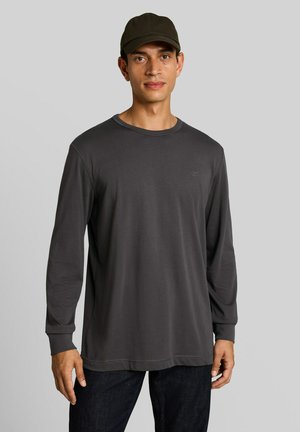 Hombre con una camisa de manga larga gris oscuro, pantalones negros y una gorra negra de pie frente a un fondo liso de color gris claro.