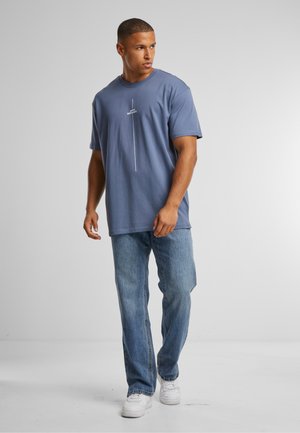 Blaue Baumwoll-T-Shirt mit kurzen Ärmeln, mit einem vertikalen weißen Grafikdesign in der Mitte. Getragen mit hellblauen Jeans und weißen Sneakers.