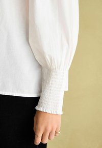 Weiße Leinenbluse mit voluminösen Ärmeln und taillierten, gerafften Bündchen. Die Bündchen sind mit zarten Rüschendetails verziert. Schwarzer Stoff ist darunter sichtbar.