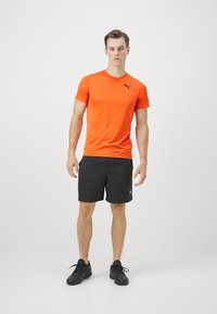 Oranžs sportisks T-krekls ar īsām piedurknēm un melnu Puma logo, apvienots ar melniem šortiem un melnām sporta kurpēm, stāvot pret vienkāršu fona.
