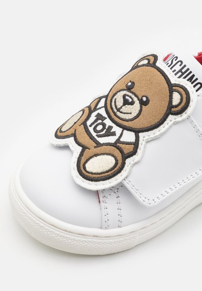 Hvit lær sneaker med en borrelåsrem og en brodert brun teddybjørn-patch med teksten "Toy". Gummisåle med teksturert mønster.