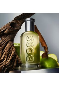Szklana butelka perfum z zielonym płynem, wytłoczone logo "BOSS", srebrna zakrętka, otoczona zielonymi jabłkami i teksturowanym drewnem.