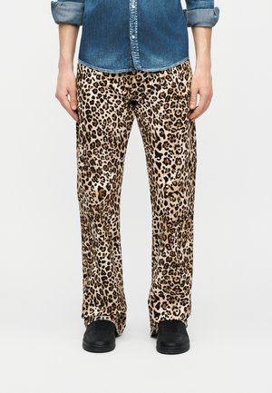 Személy leopárdmintás nadrágban, fekete cipőben és kék farmeringben, felhajtott ujjakkal, egyszínű fehér háttér előtt.