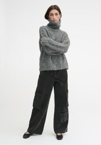 Siva pletena pulover s turtleneck in ohlapnim krojem, v kombinaciji z velikimi črnimi cargo hlačami z velikimi žepi in ravnimi črnimi čevlji.