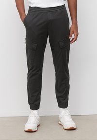 Černé cargo kalhoty s pružným stahováním v pase a elastickými manžetami, se dvěma bočními kapsami a hladkým, lehkým materiálem. Nosí se s bílými teniskami.