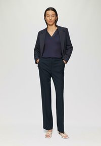 Blazer navy avec des épaules structurées, associé à un haut navy à décolleté en V et un pantalon sombre à motif. Accentué par des sandales à talons blanches.