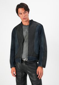 Denim jaka ar tumši zilo un melno paneļu kombināciju, ar rāvējslēdzēju un stāvošu apkakli. Valkāta uz pelēka grafiskā T-kreklu.