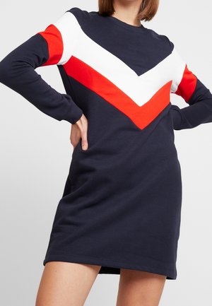 Frau trägt ein marineblaues Kleid mit weißen und roten Chevron-Streifen, Hände in Hüfthöhe, vor einem schlichten hellgrauen Hintergrund.