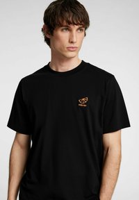 Svart bomullströja med en liten orange broderad logotyp på vänster bröst. Korta ärmar och rundad halsdesign.