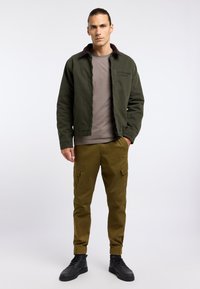 Green canvas jacket with a brown corduroy collar, paired with olive cargo pants and black lace-up boots. Textured fabric, fitted design.