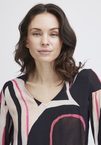 Blouse fluide à col en V noir, ornée de motifs abstraits en rose, blanc et marron. Tissu léger avec des manches longues et une coupe décontractée.