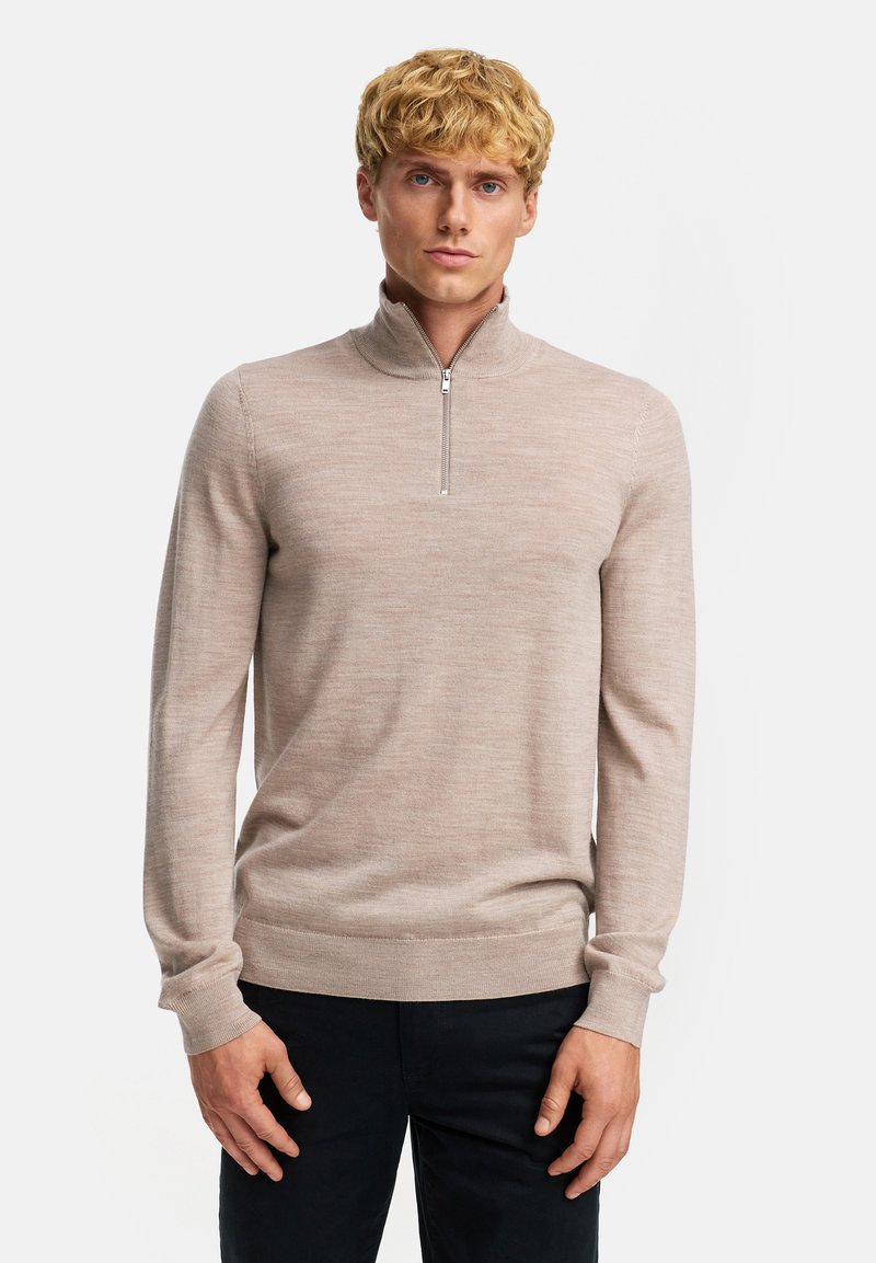 Blond man som bär en beige långärmad tröja med halvdragkedja och svarta byxor, stående mot en vit bakgrund.