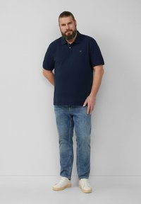 Granatowa koszulka polo, krótkie rękawy, miękka bawełniana tkanina, noszona z jasnoniebieskimi dżinsami i białymi trampkami, stojąca na neutralnym tle.