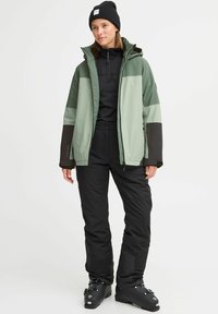 Grüne und schwarze Skijacke mit Reißverschlusstaschen, getragen über einem schwarzen Oberteil und einer Hose. Mit Kapuze und strukturiertem Stoff. Schwarze Mütze inklusive.