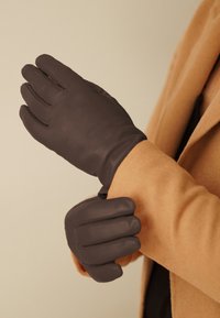 Gants en cuir marron avec une texture lisse et un design à cinq doigts, portés sur une main contre un fond clair et un manteau beige.