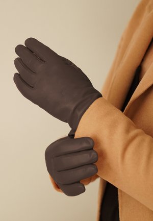 Braune Lederhandschuhe mit glatter Textur und fünf Fingerdesign, getragen an einer Hand vor einem hellen Hintergrund und einem beigen Mantel.