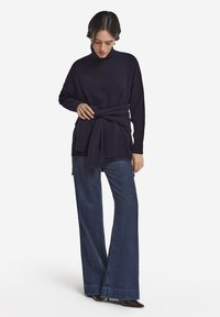 Marinblå stickad polotröja med skärp i midjan, kombinerad med vida jeansbyxor och spetsiga skor, med en ribbad textur.