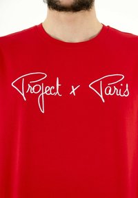 Red cotton t-shirt featuring white cursive text "Project x Paris" across the chest. Smooth texture with a classic crew neck design.