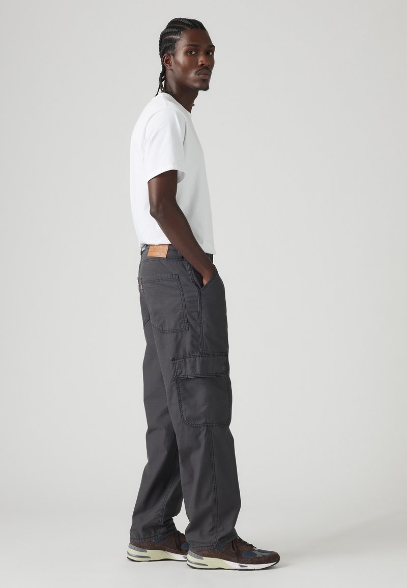 Pantalones cargo grises con múltiples bolsillos y un parche de cuero en la cintura, combinados con una camiseta blanca sencilla y zapatillas oscuras.