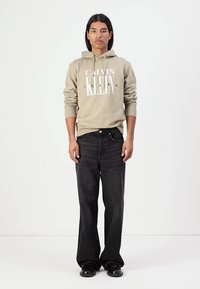 Sudadera con capucha color caqui con texto blanco y jeans negros, ambos de corte casual. El modelo está de pie, con zapatos negros, sobre un fondo simple.