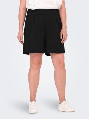 Kvinde iført sorte knælange shorts, hvid blondetop og hvide sneakers står mod en ensfarvet baggrund.