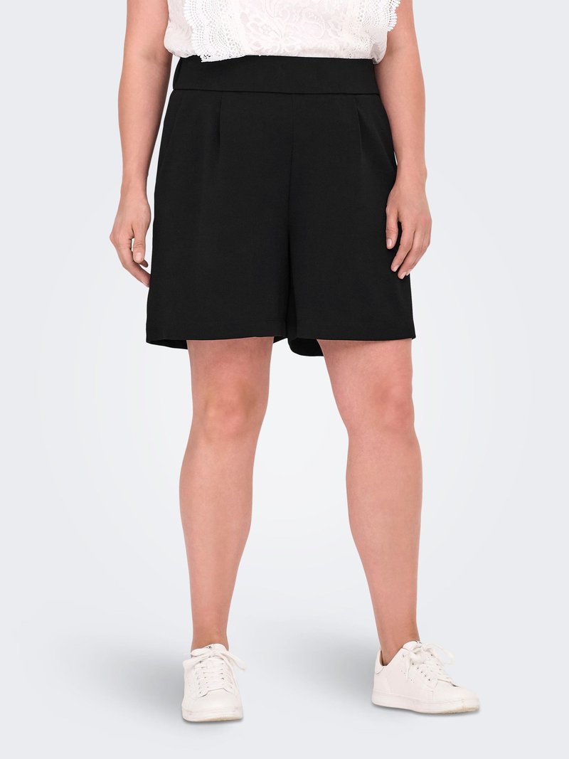 Mujer con pantalones cortos negros hasta la rodilla, top blanco de encaje y zapatillas blancas, de pie contra un fondo liso.