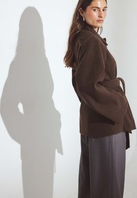 Hnedý sak s pásom a širokými rukávmi, so zavinovacím dizajnom a textúrovaným povrchom. V kombinácii s tmavými nohavicami, vrhajúcimi jemný tieň.