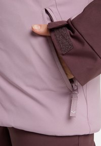 Paarse jas met een zachte textuur, voorzien van een zijzak en klittenbandsluiting. Inclusief een ritssluiting met een hartaccent.