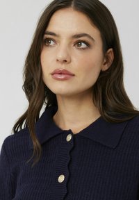 Pull en maille côtelée bleu marine avec une patte de boutonnage à festons et des boutons dorés, doté d'un col classique et d'un tissu texturé.