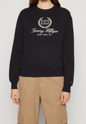 Osoba nosząca czarną bluzę Tommy Hilfiger z logo i napisem "New York, NY", połączoną z beżowymi spodniami cargo.