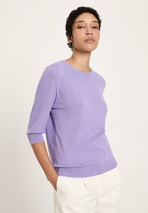 Frau mit kurzen, lockigen Haaren, die einen gerippten hellvioletten Pullover mit Dreiviertelärmeln und weiße Hosen trägt, stehend mit Händen in den Taschen.