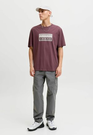 Mand med hvid kasket, vinrød Jack & Jones t-shirt, grå cargobukser, hvide sneakers og en sølvkæde, står mod en ensfarvet baggrund.