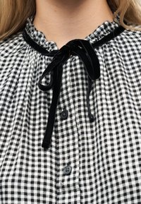 Gros plan sur un chemisier à carreaux noir et blanc avec un col à volants et un nœud en velours noir noué au niveau de l'encolure.