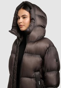 Kohev pruun alla jope kõrge krae ja kapuutsiga. Tekstureeritud, läikiv kangas, nähtavate õmblustega ja küljetasku detailiga.