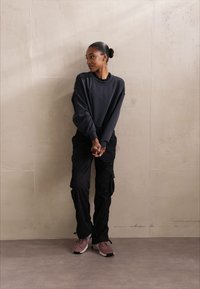 Fekete, oversize pulóver, fekete cargo nadrág és rózsaszín sportcipő. Simább anyag, laza fazon és praktikus zsebek. Semleges háttér előtt stílusosan elhelyezve.