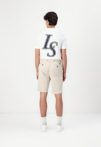 Witte t-shirt met een grote bedrukte "IS" op de achterkant, gecombineerd met beige shorts en witte sneakers. Eenvoudig ontwerp en neutrale kleuren.