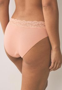 Kobieta w brzoskwiniowej bieliźnie ozdobionej koronką, widziana od tyłu, z jedną ręką swobodnie opuszczoną wzdłuż ciała.