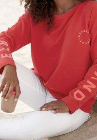 Camicia rossa a maniche lunghe con scritte bianche sulle maniche, abbinata a pantaloni bianchi. Persona che tiene in mano una tazza con coperchio rosa, seduta sulla sabbia.