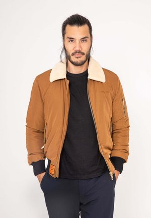 Brown bomber jacket with a beige faux fur collar, zippered pocket details, ribbed cuffs, and an orange label on the left side.
