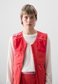 Rote ärmellose Weste mit Frontreißverschluss, mehreren Taschen, kontrastierenden pinken Reißverschlussakzenten, getragen über einem weißen langärmligen Shirt.
