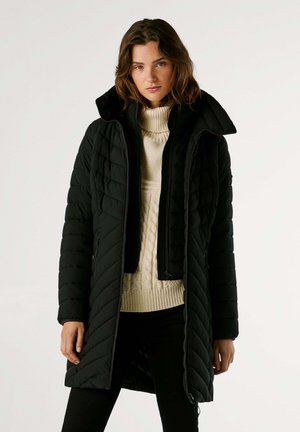 Cappotto imbottito nero trapuntato con colletto alto e chiusura a zip, abbinato a un maglione a trecce color crema e pantaloni scuri. Superficie testurizzata.