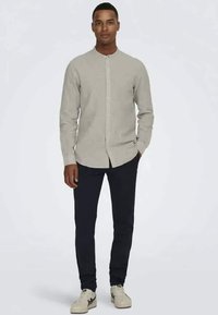 Camisa cinza claro, com colarinho redondo, combinada com calças slim-fit escuras e ténis bege, de pé contra um fundo simples.