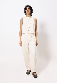 Lichtbeige, cropped mouwloos topje met vijf gouden knopen, gecombineerd met bijpassende textuur brede broek en zwarte gestreepte flats.