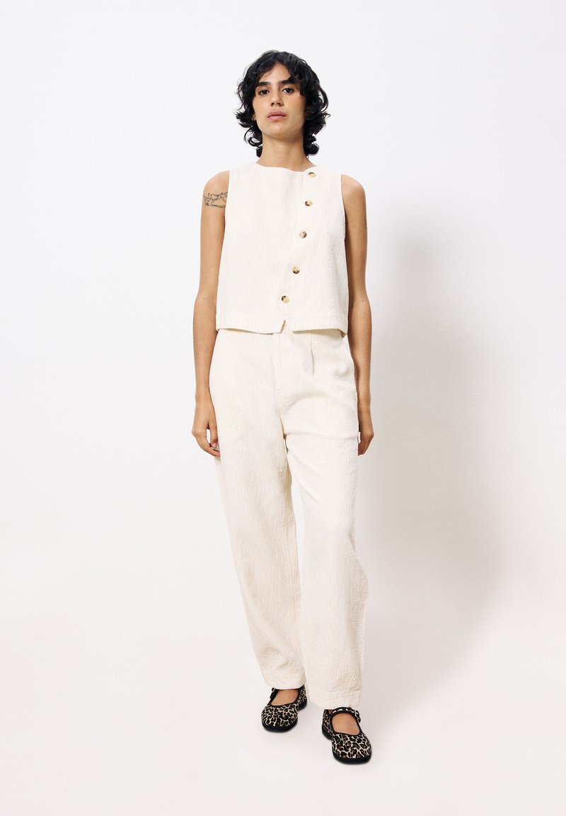 Lichtbeige, cropped mouwloos topje met vijf gouden knopen, gecombineerd met bijpassende textuur brede broek en zwarte gestreepte flats.