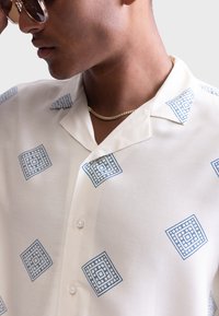 Chemise à manches courtes en crème avec des motifs géométriques bleus. Tissu léger avec une texture lisse, dotée d'un col et d'une fermeture à boutons.
