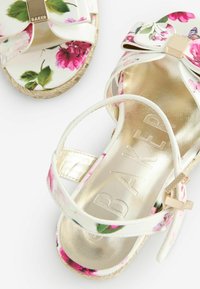 I sandali con stampa floreale presentano una parte superiore bianca lucida, dettagli dorati e una suola in stile espadrilles con una trama strutturata. Fiori bianchi e multicolori.