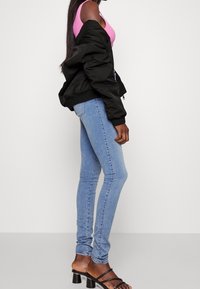 Femme portant un jean skinny bleu clair, des sandales noires à talons, un top rose sans manches et une veste noire tombant sur ses épaules.