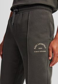 Pantaloni de trening de culoare verde măsliniu închis, realizați dintr-un material moale. Au talie elastică, șnur de strângere și un logo brodat pe buzunar.