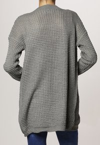 Back view of a person wearing a loose, long gray waffle-knit cardigan over dark pants against a plain background.