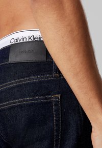 Jeans de mezclilla azul oscuro con un parche de cuero negro en la cintura. Presenta costuras en contraste y una etiqueta blanca visible en la cintura.